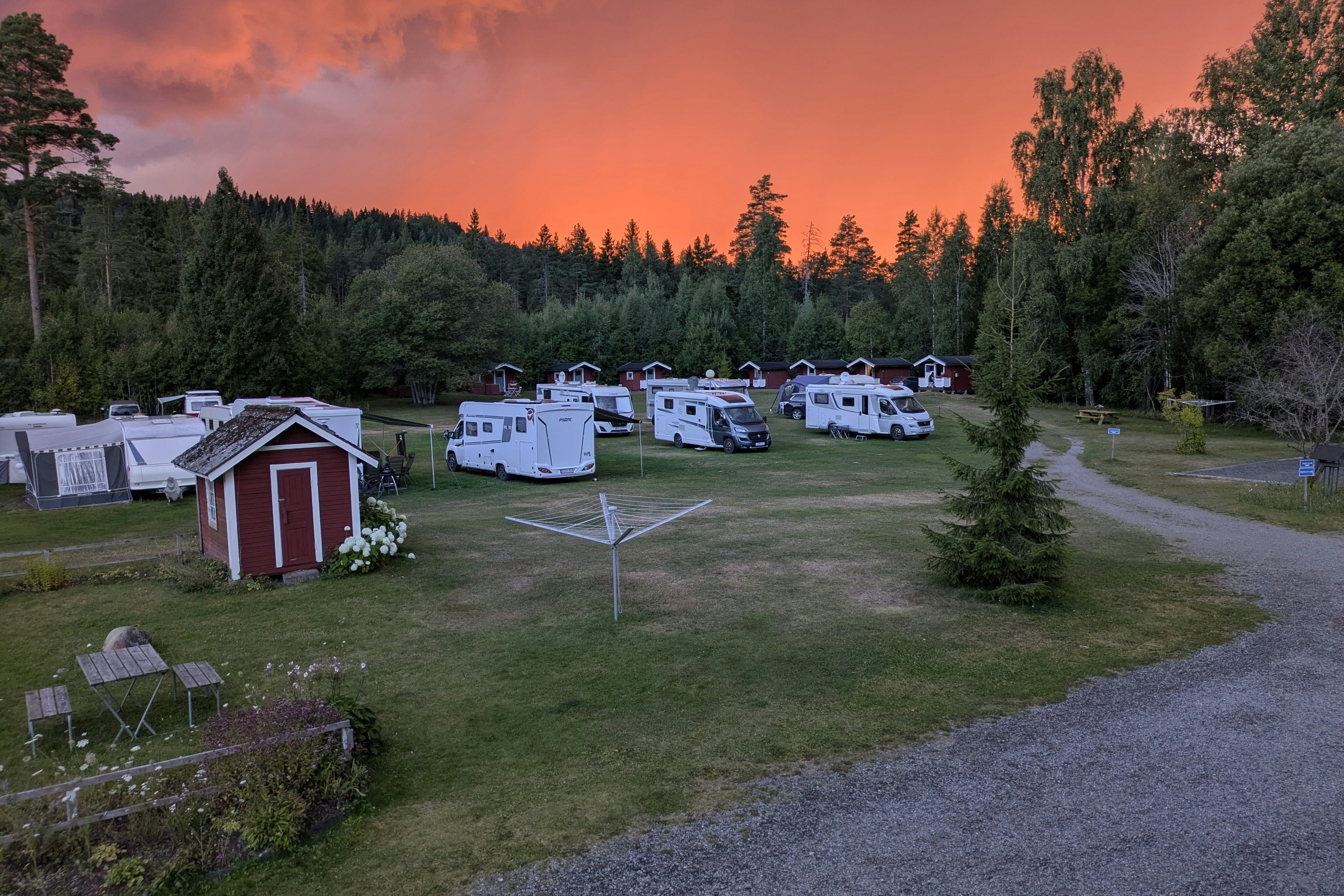 Campingplatz mit mehreren Wohnwagen und Stellplätzen, Rasenfläche, Wegen und einem kleinen roten Geräteschuppen im Abendlicht.