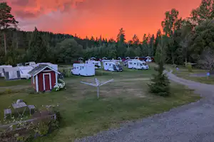 Campingplatz mit mehreren Wohnwagen und Stellplätzen, Rasenfläche, Wegen und einem kleinen roten Geräteschuppen im Abendlicht.