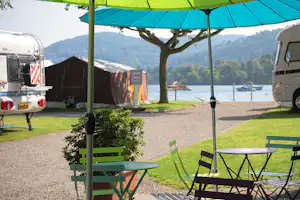 Campingplatz mit Stellplätzen, Wohnwagen und Zelt unter Sonnenschirm, Kiesweg und Blick auf den See zwischen Bäumen.