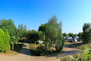 Campingplatz mit gepflasterten Wegen, Hecken und Bäumen, im Hintergrund Wohnwagen und ein Wohnmobilstellplatz.