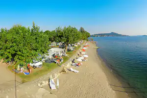 Campingplatz am Seeufer mit Strand, Liegen und Bäumen; Wohnwagen und Autos stehen entlang des Weges.
