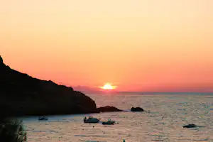 Sonnenuntergang über dem Meer mit felsiger Bucht, ruhigem Wasser und kleinen Booten am Ufer.