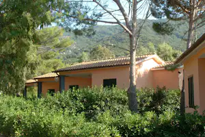 Campinggebäude mit gelber Fassade, Veranda und Hecken am Weg, umgeben von Bäumen und Hügeln im Hintergrund.