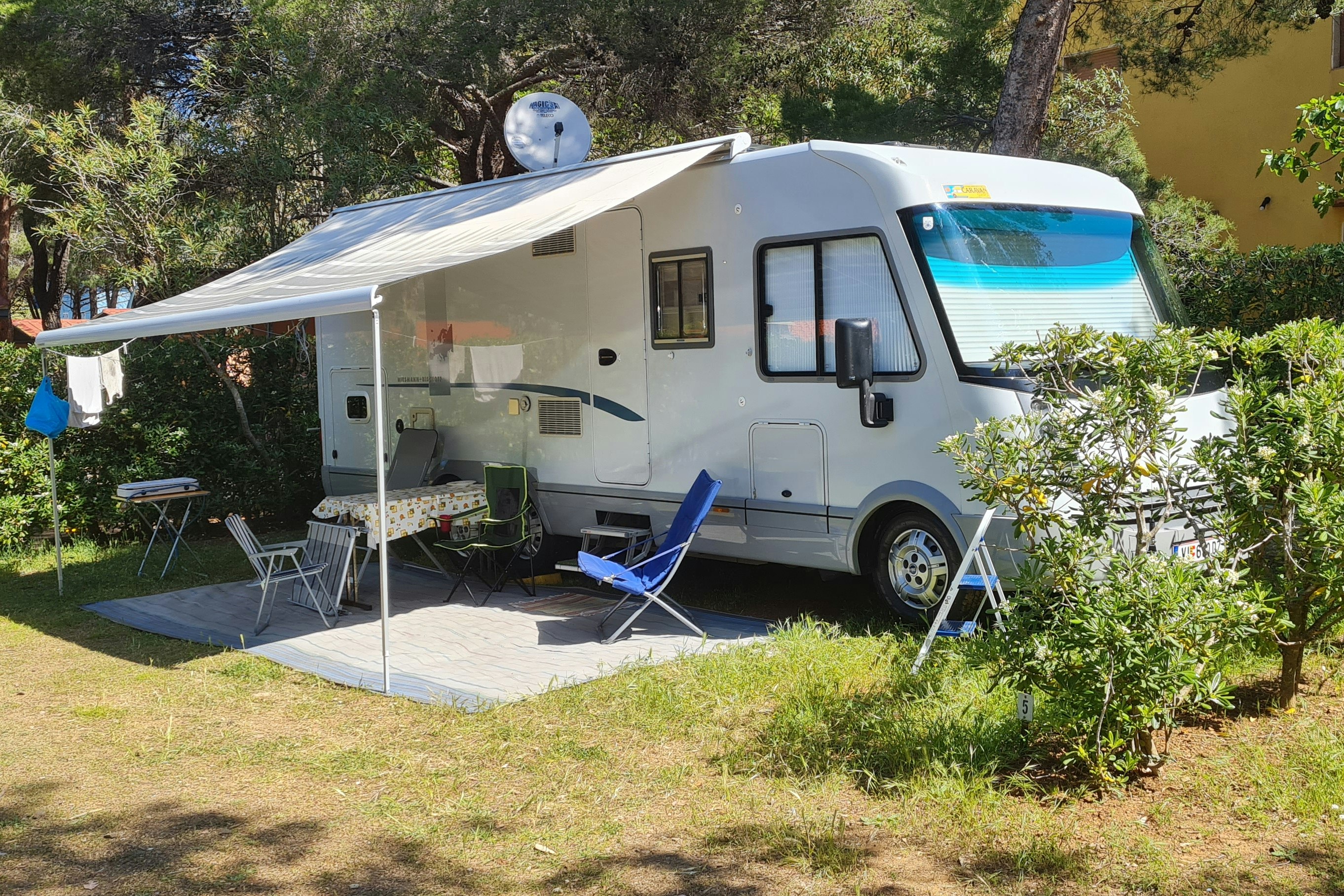 Wohnmobil mit Vorzelt und Sitzbereich auf einem Campingplatz im Grünen, umgeben von Bäumen und Rasen.