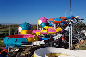 Bunte Wasserrutschen auf einem Spielplatz im Freien, mit Stahlgeländer und Blick auf Wiesen und Bäume.
