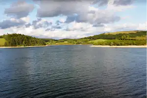Blick auf einen See mit bewaldeten Ufern, grünen Hügeln und Wolken am Himmel.