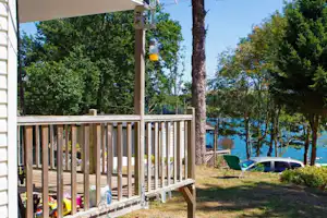 Holzveranda am Wohnwagen-Stellplatz, daneben Liegen und Rasenfläche mit Bäumen und Blick auf den Fluss.