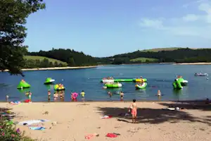 Sandstrand am See mit aufblasbaren Booten und Schwimmreifen im Wasser, daneben liegen Handtücher und Badekleidung.