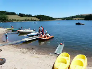 Bootssteg am See mit mehreren Booten, gelben Schwimmhilfen und Personen am Ufer, im Hintergrund grüne Hügel.