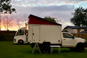 Wohnmobil auf einem Stellplatz am Campingplatz, mit Leiter und Tisch, umgeben von Bäumen und Abendhimmel.