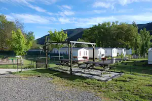 Holzterrasse mit Picknicktischen auf einem Campingplatz, im Hintergrund Reihen von weißen Mobilheimen und grüne Bäume.