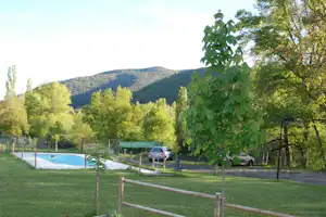 Grüner Campingplatz mit Pool, Auto am Stellplatz und Bäumen vor bewaldeten Hügeln im Hintergrund.