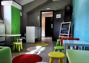 Flur in einem Campinggebäude mit bunten Sitzhockern, Tisch und grünen Türen, daneben ein Mülleimer und ein Eingang.