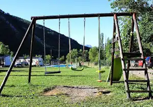Schaukeln auf einem grünen Camping-Spielplatz, im Hintergrund Bäume, ein Pool und Berge.