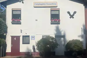 Gebäude am Campingplatz Sonnenberg mit Eingang, zwei Fenstern mit Markisen und Zierpflanzen vor der Fassade.