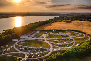 Luftaufnahme eines Campingplatzes am See mit Stellplätzen, Wohnmobilen, Gebäuden und Zufahrtswegen im Abendlicht.