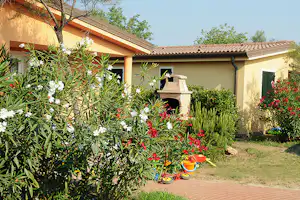 Bungalow mit Terrasse und Blumenbeet, im Vordergrund Sträucher und bunte Blüten vor dem gelben Gebäude.