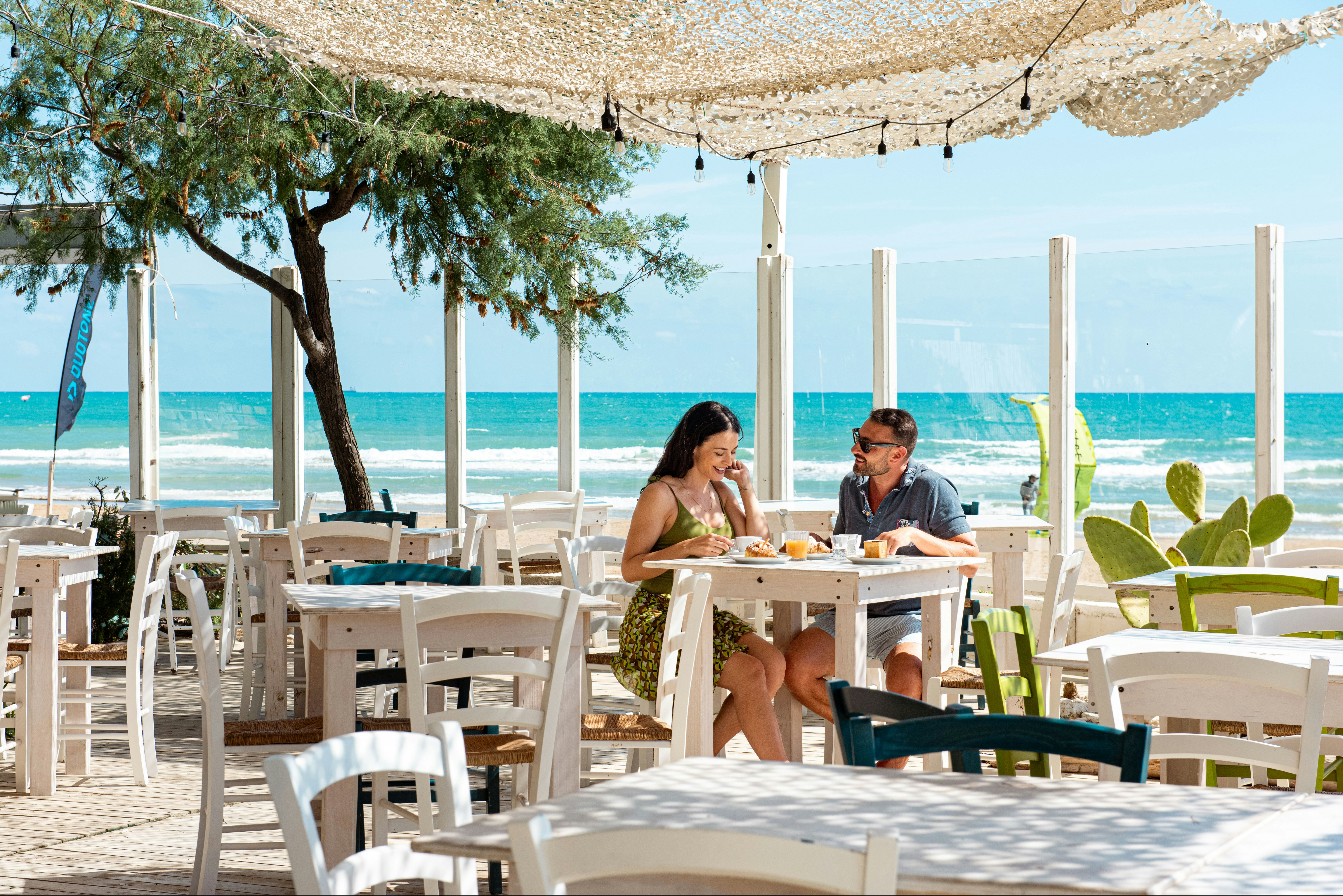 Weißes Restaurant-Set mit Tischen und Stühlen unter Sonnensegeln, zwei Personen essen am Meer mit Blick auf das Wasser.