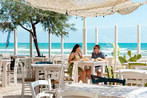 Weißes Restaurant-Set mit Tischen und Stühlen unter Sonnensegeln, zwei Personen essen am Meer mit Blick auf das Wasser.