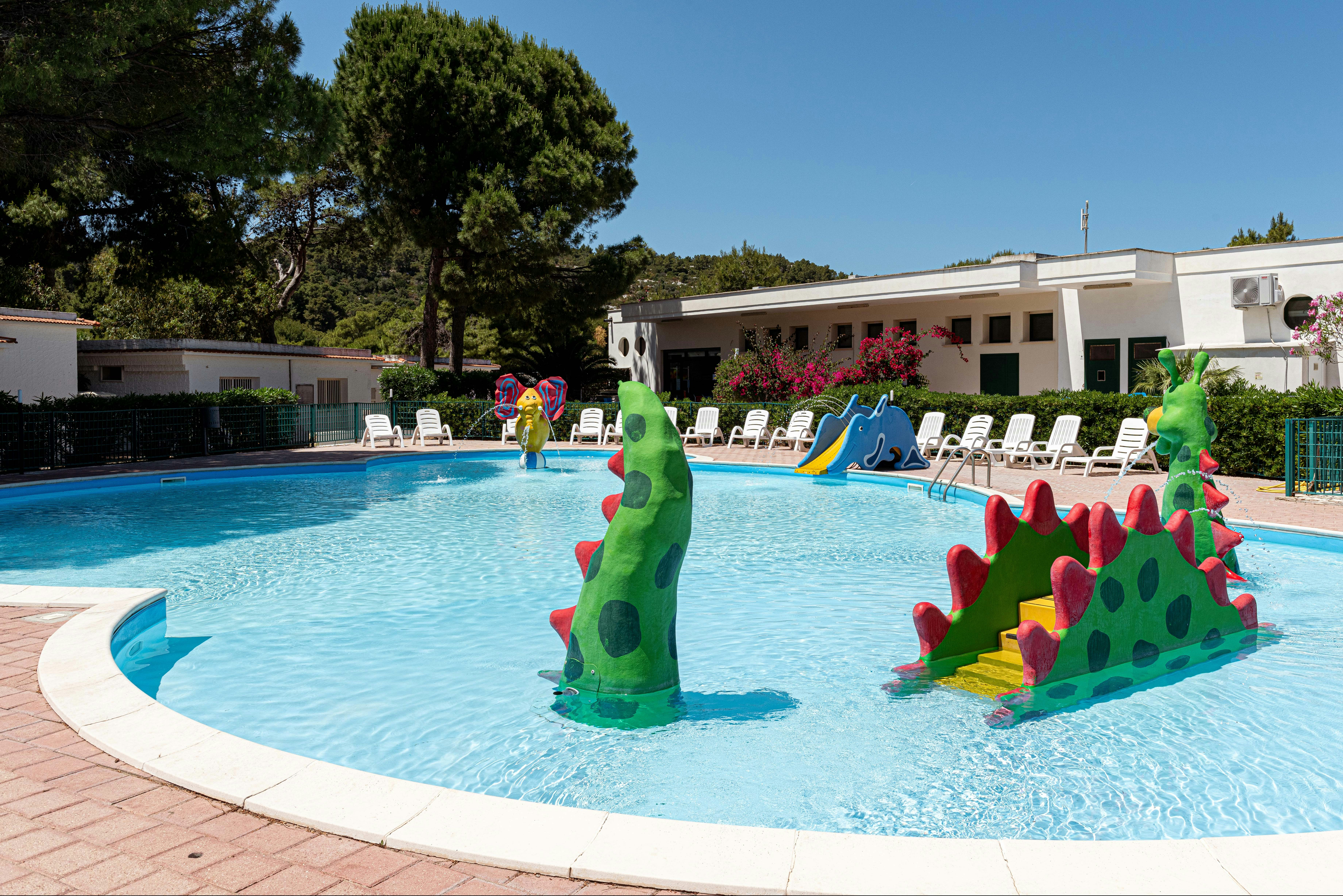 Schwimmbecken mit bunten Wasserspiel-Figuren, Liegen am Rand und weißen Gebäuden im Hintergrund, umgeben von Bäumen.