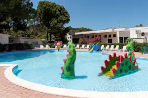 Schwimmbecken mit bunten Wasserspiel-Figuren, Liegen am Rand und weißen Gebäuden im Hintergrund, umgeben von Bäumen.