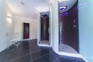 Badezimmer im Campingbereich mit offener Glastür, Dusche mit Mosaikfliesen und beleuchtetem Duschkopf, daneben Stuhl und Fliesenboden.