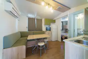 Wohnbereich mit Sofa, Holztisch und Küchenzeile in einer Campingunterkunft, mit Vorhängen, TV und Kühlschrank im Hintergrund.