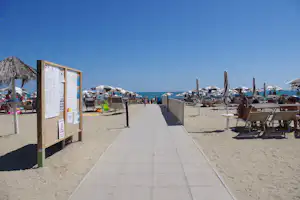Weg am Strand mit Werbetafel, Liegestühlen und Sonnenschirmen, im Hintergrund das Meer unter blauem Himmel.