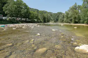 Fluss mit klarer Strömung, Kiesel und Steinen, im Hintergrund Bäume und ein Campingbereich mit Wohnwagen.