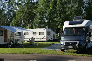 Wohnmobil und Wohnwagen auf dem Campingplatz, davor ein Sonnensegel und Campingstühle auf dem Rasen, im Hintergrund Bäume.