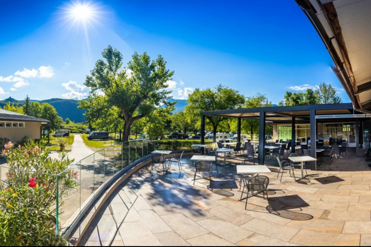 Terrasse mit Sitzgarnituren unter einem überdachten Bereich, gepflasterter Weg und Bäume im Campingbereich vor grünen Hügeln.