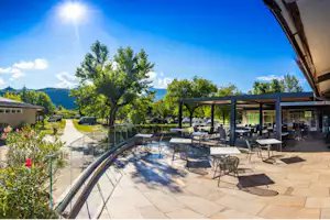 Terrasse mit Sitzgarnituren unter einem überdachten Bereich, gepflasterter Weg und Bäume im Campingbereich vor grünen Hügeln.