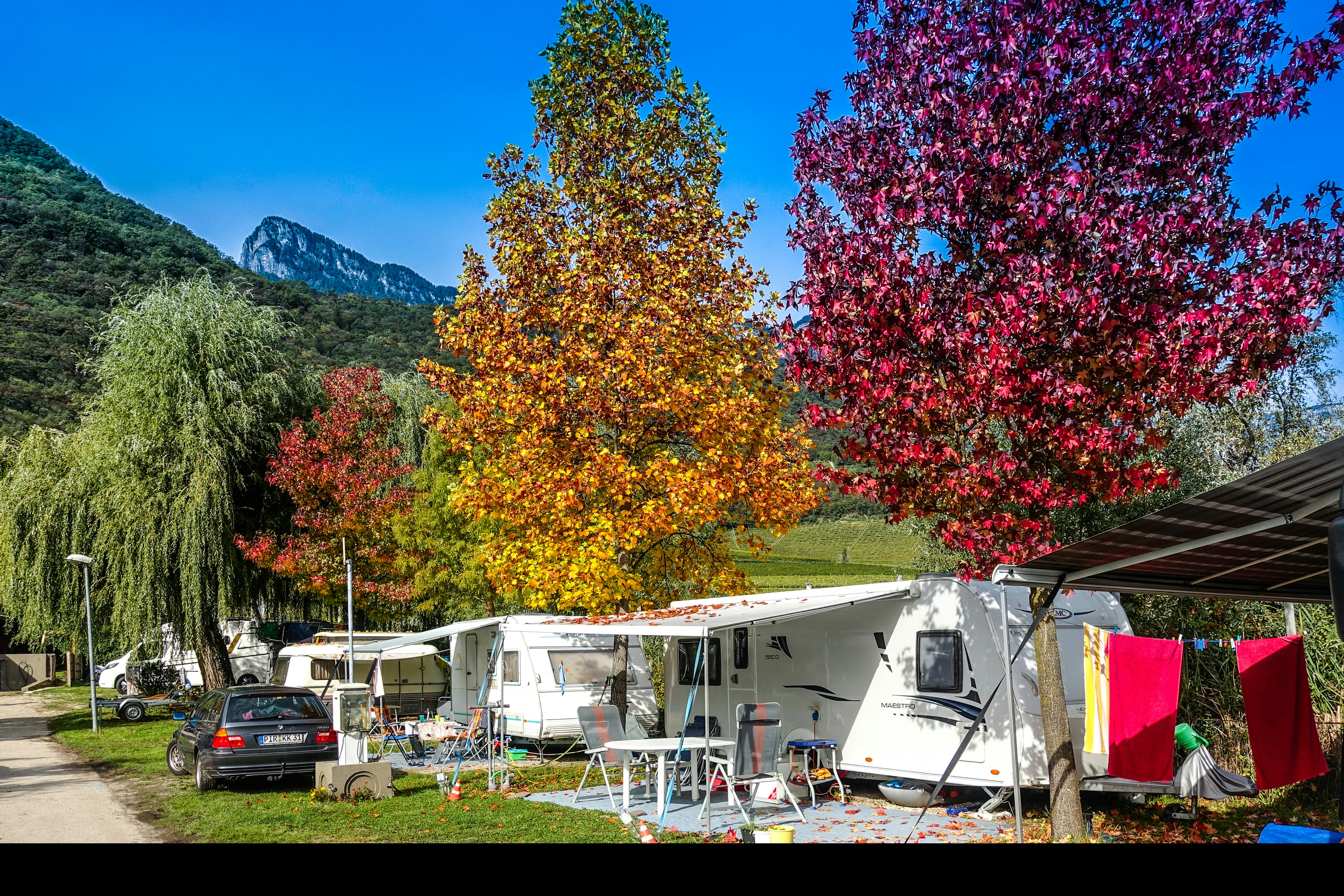 Campingplatz mit Wohnwagen und Zelten, Wäscheleine und Markise am Stellplatz, umgeben von bunten Bäumen und Bergen im Hintergrund.