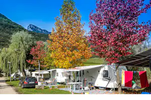 Campingplatz mit Wohnwagen und Zelten, Wäscheleine und Markise am Stellplatz, umgeben von bunten Bäumen und Bergen im Hintergrund.
