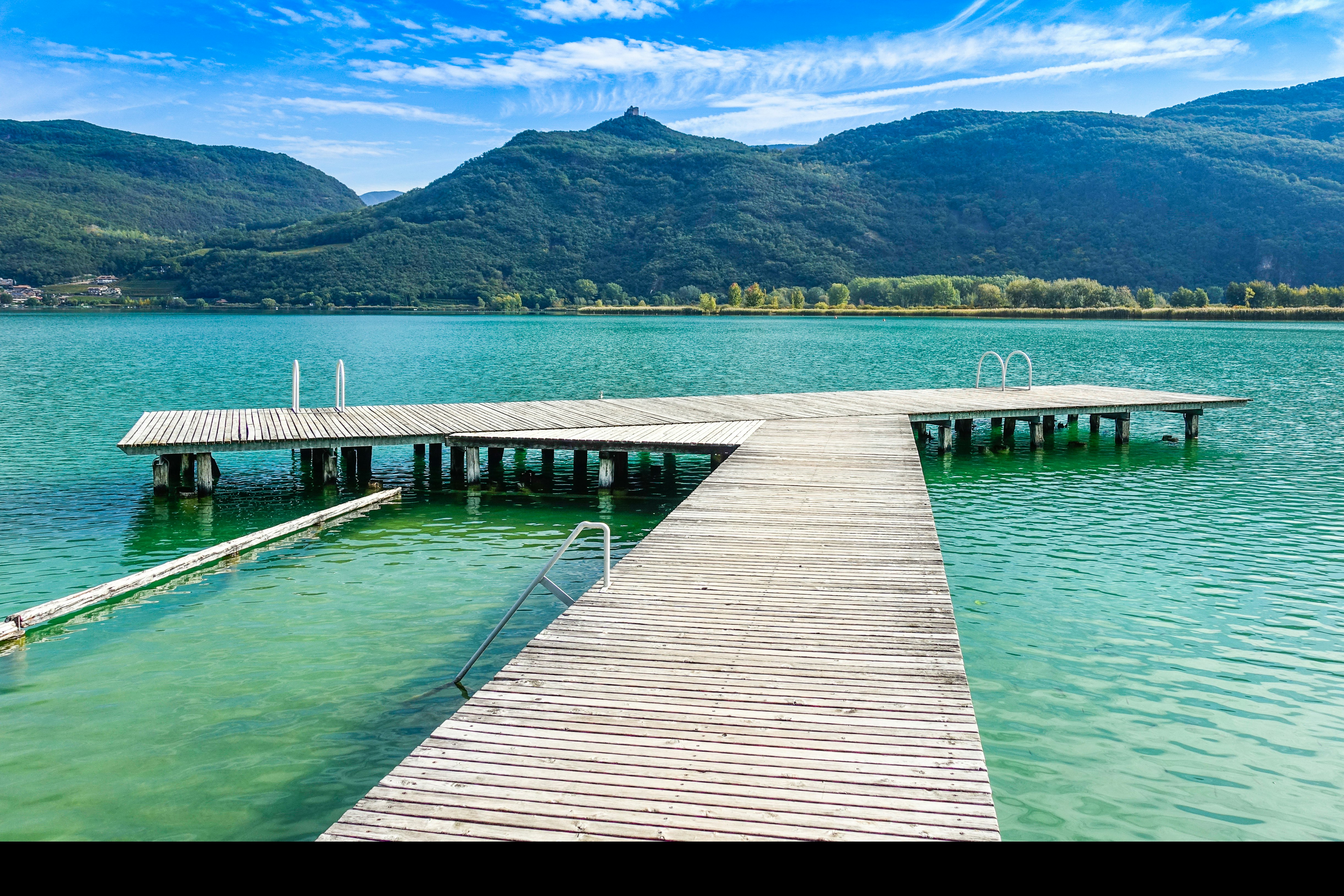 Holzsteg am See mit mehreren Bootsstegen und Pollern, im Hintergrund grüne Hügel und Berge unter klarem Himmel.