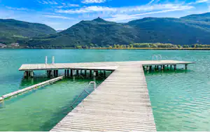 Holzsteg am See mit mehreren Bootsstegen und Pollern, im Hintergrund grüne Hügel und Berge unter klarem Himmel.