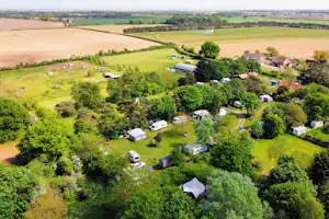 Luftaufnahme eines Campingplatzes mit Wohnmobilen, Zelten und einem großen Zelt auf grüner Wiese zwischen Feldern und Bäumen.