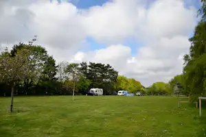 Campingplatz mit Wohnwagen und Campern auf grünem Rasen, umgeben von Bäumen und einem Zaun.