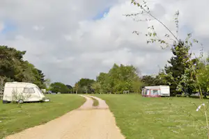 Wohnwagen und Zelt auf einem grünen Campingplatz, Kiesweg zwischen den Stellplätzen, Bäume und Himmel mit Wolken.