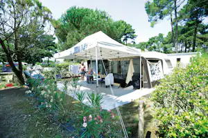 Campingzelt mit Vorzelt auf einem Stellplatz im Grünen, mit Tisch und Stühlen unter dem Dach und Bäumen im Hintergrund.