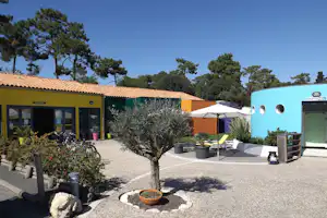 Campinganlage mit Kiesweg, Olivenbaum im Vordergrund, Sitzbereich mit Sonnenschirm und Sanitärgebäude mit Beschriftung „Toilettes“.