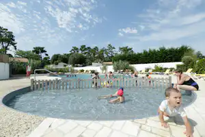 Schwimmbecken auf dem Campingplatz mit Kinderbereich, weißen Liegen und Sonnenschirmen, umgeben von Bäumen und Hecken.