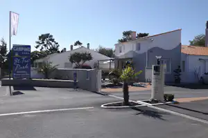 Campingplatz-Eingang mit Schildern, weißem Gebäude und Rezeption, davor Hecken, Palmen und ein Parkplatz am Straßenrand.