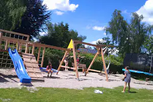 Spielplatz auf dem Campingplatz: Rutsche und Schaukel auf dem Rasen, Holzkonstruktionen und Kinder im Hintergrund.