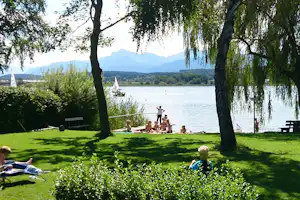 Grüner Campingplatz am See mit Liegestühlen, Picknicktisch und Bäumen am Ufer, im Hintergrund Berge und Wasser.