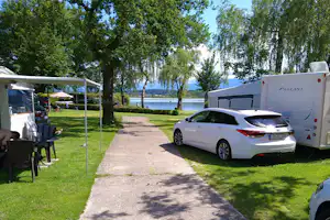 Stellplatz auf dem Campingplatz mit Wohnwagen und Auto auf der Wiese, daneben Schatten von Bäumen und ein Tisch unter Markise.