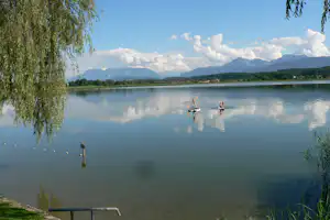 Ruhiger See mit Spiegelung, Ruderboot im Wasser und Blick auf Berge; am Ufer grünes Gras und Bäume.