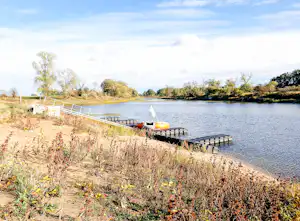 Ufer am See mit Steg und Boot am Campingplatz, Gras und Sträucher am Ufer, ruhiges Wasser im Hintergrund.
