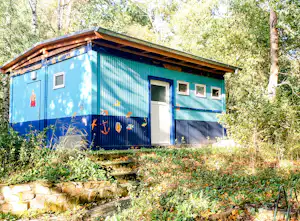 Blauer Bungalow im Wald mit Holzveranda, Fenstern und Eingangstür, umgeben von Büschen und Bäumen.