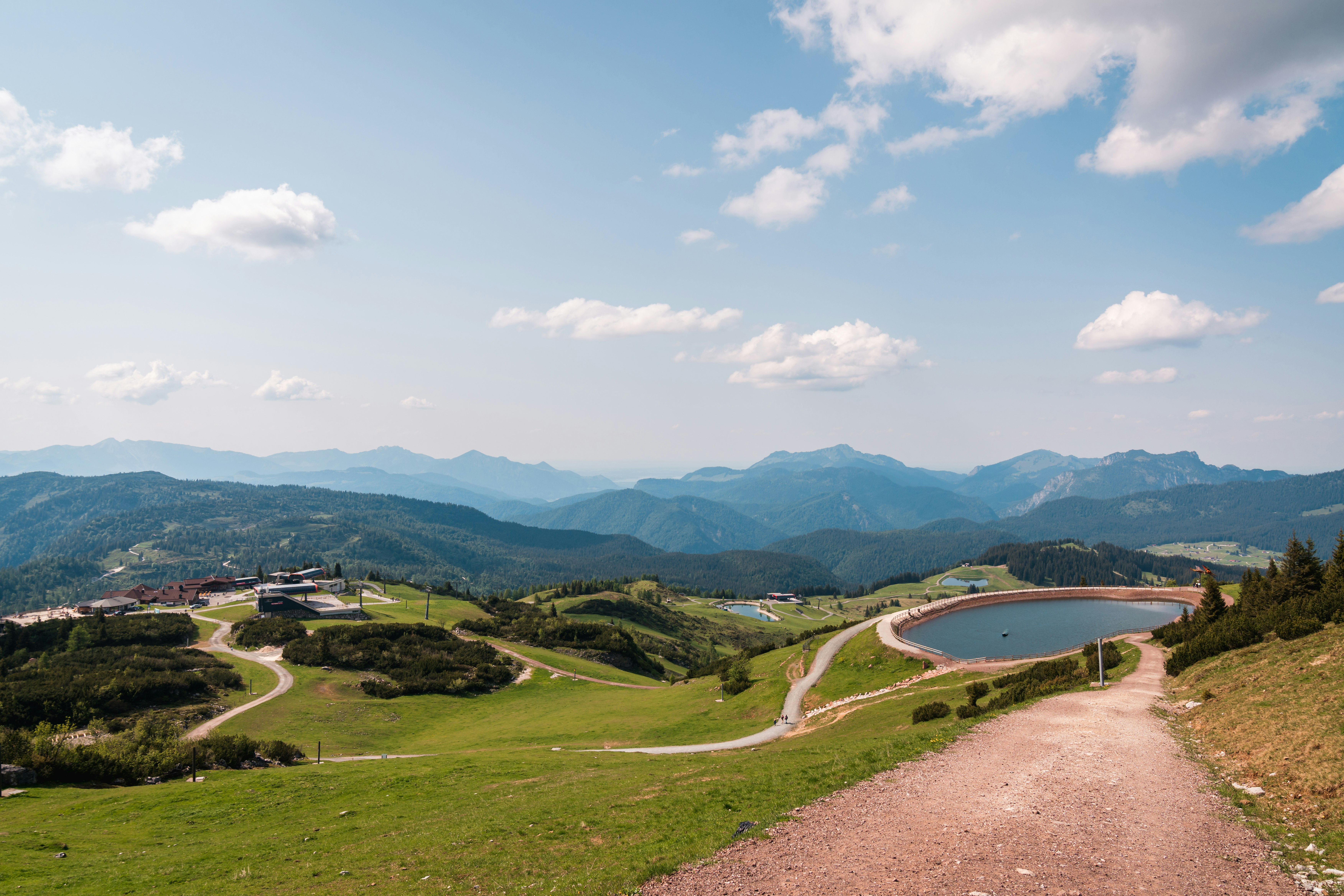 Campinganlage mit See, Uferweg und Wohngebäuden im Tal, umgeben von grünen Wiesen und Bergen unter blauem Himmel.